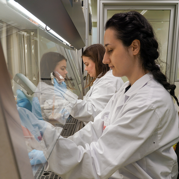 Biologisches Labor mit zwei Personen von der Seite aufgenommen, die vor eine Abzugshaube im weißen Laborkittel sitzen und Flüssigkeiten pipetieren.